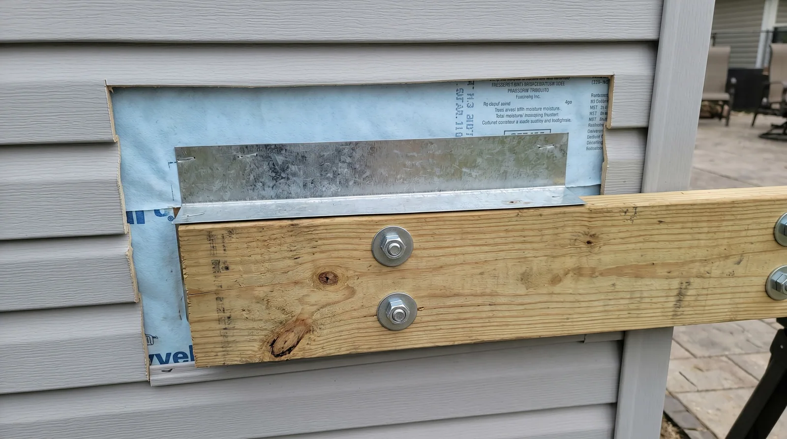 Close-up of a deck ledger flashing installation on a vinyl-sided home, showing the flashing pan and bolts, with siding removed.