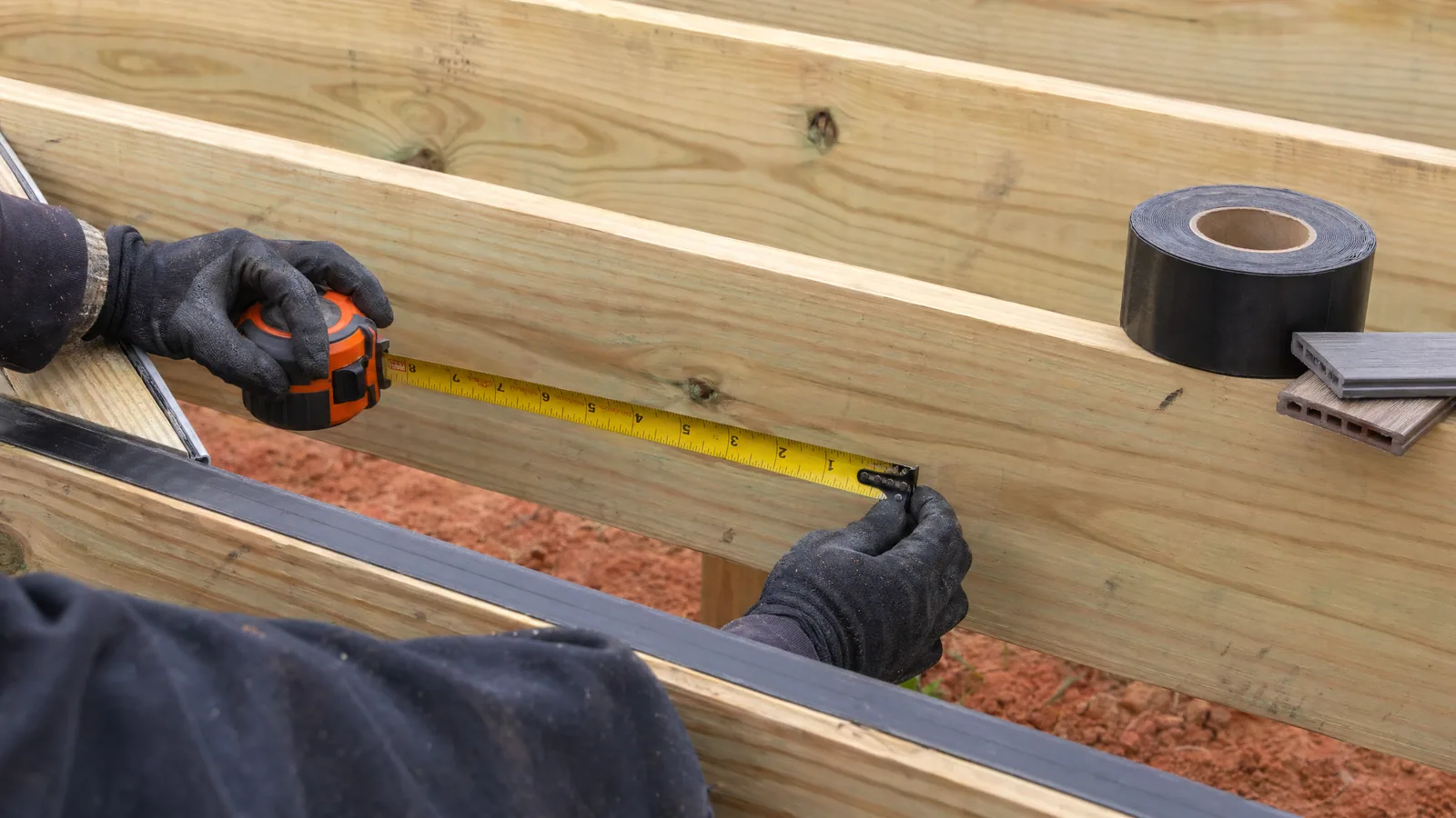 Deck builder measuring joist spacing on a Raleigh jobsite with joist tape and composite decking samples over Piedmont clay soil