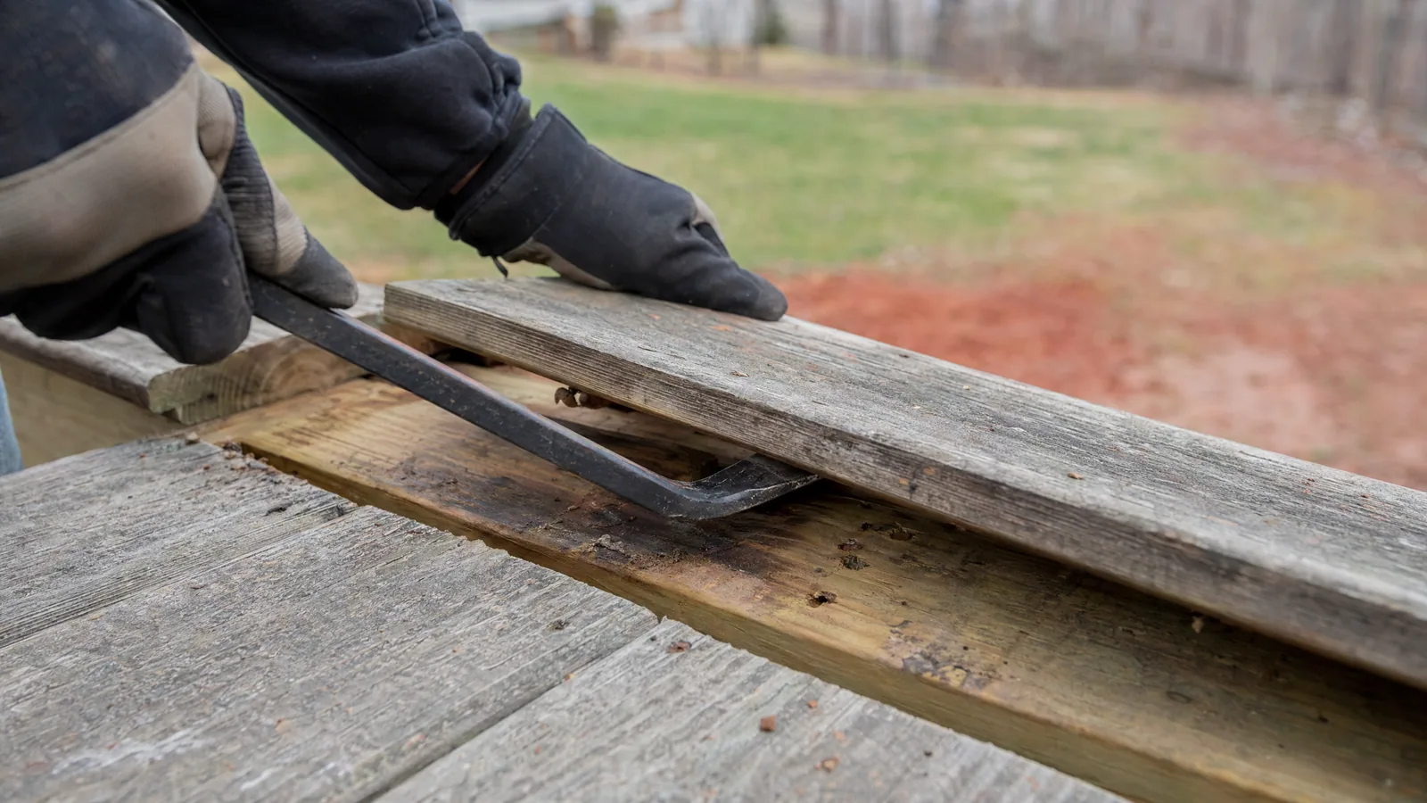 Close-up of a contractor prying up an old deck board to inspect the condition of the underlying joist during a resurfacing evaluation.